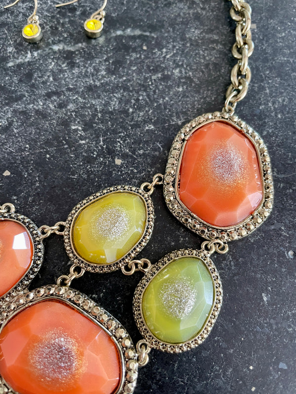 A close-up of a statement necklace with irregular orange and yellow-green cabochon stones in ornate silver-tone settings, shown on a dark textured surface with matching drop earrings partially visible at the top left.