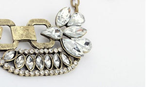 A close-up of a statement necklace featuring a textured antique gold-tone chain and a large floral cluster of clear teardrop rhinestones on a white background.