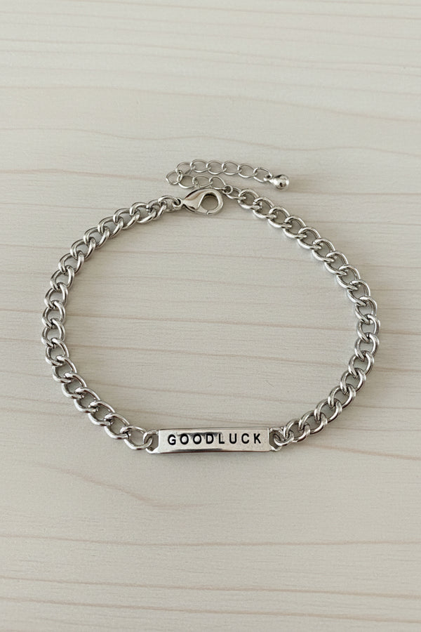 A silver chain bracelet with a rectangular bar engraved with the word 'GOODLUCK', displayed on a textured gray stone surface.