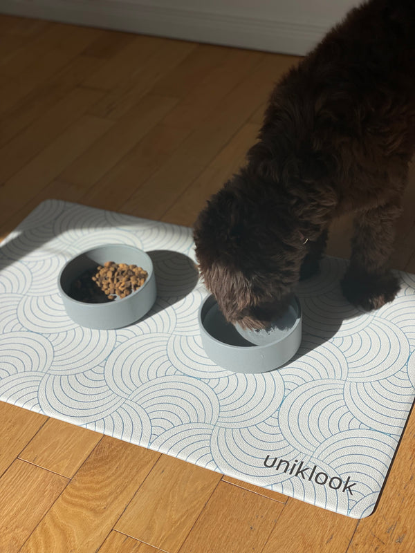 Dog eating from a bowl on a 'unikook' branded mat on a wooden floor.