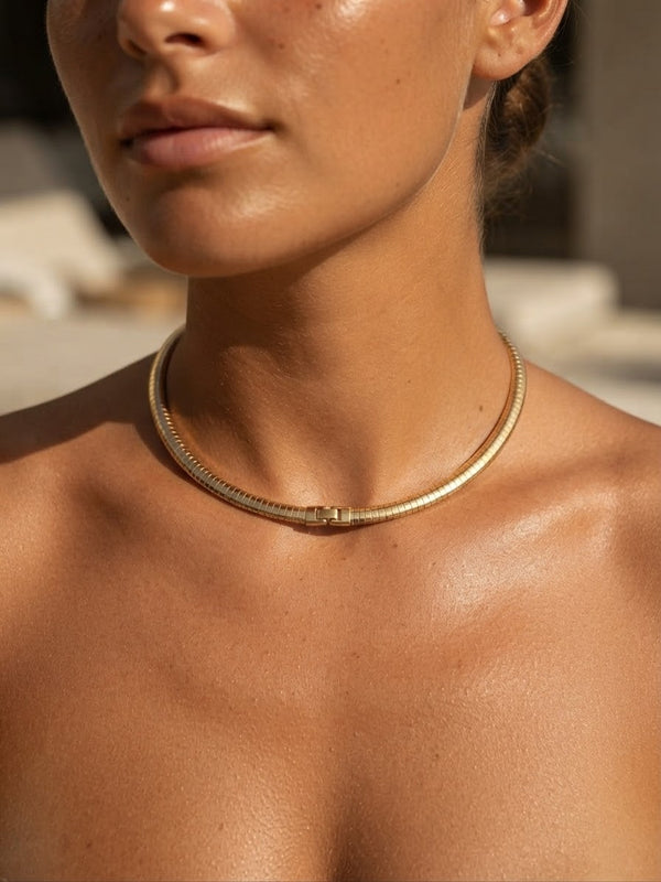 A close-up lifestyle shot of a model wearing a delicate gold snake chain necklace with a small central clasp, set against a softly blurred background.