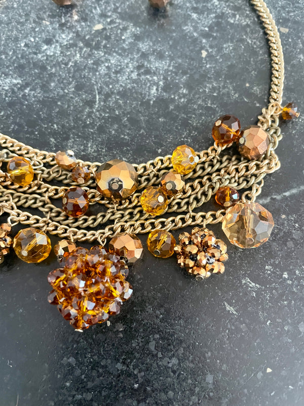 A close-up of a multi-strand statement necklace featuring antique gold-tone chains, faceted amber glass beads, and textured crystal clusters on a dark gray stone background.