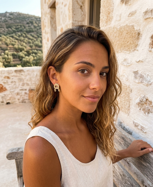 Woman with wavy hair and earrings standing outdoors near a stone wall.