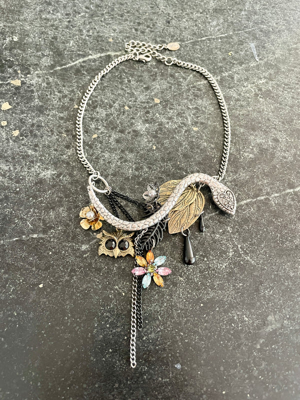 A silver-toned statement necklace featuring a snake, leaf, owl, and colorful flower charms, displayed on a dark textured surface.