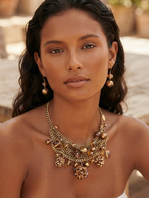 A close-up of a woman wearing a chunky gold-toned statement necklace with dangling charms and matching drop earrings, set against a softly blurred warm outdoor background.