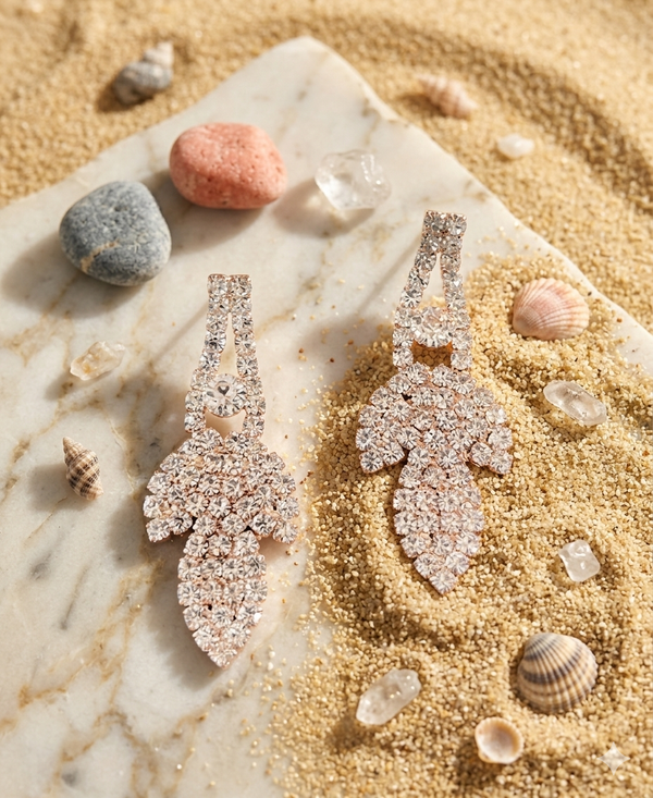 Pair of sparkling earrings on a marble surface with sand and shells around