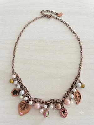 A copper-toned statement necklace with multiple dangling charms, including heart-shaped pendants, a leaf charm, flower charms, and faux pearl beads, displayed on a light wood background.