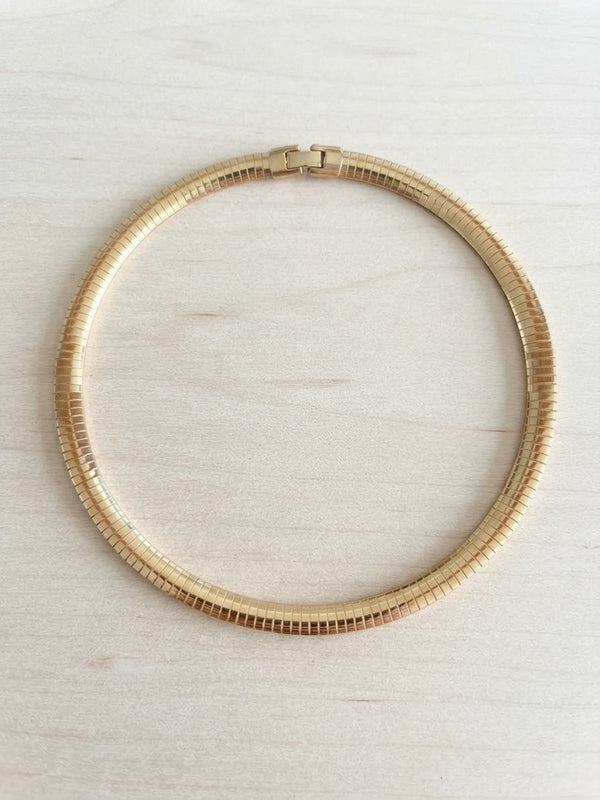 A gold-toned snake chain necklace arranged in a circle on a light beige wood-grain surface.