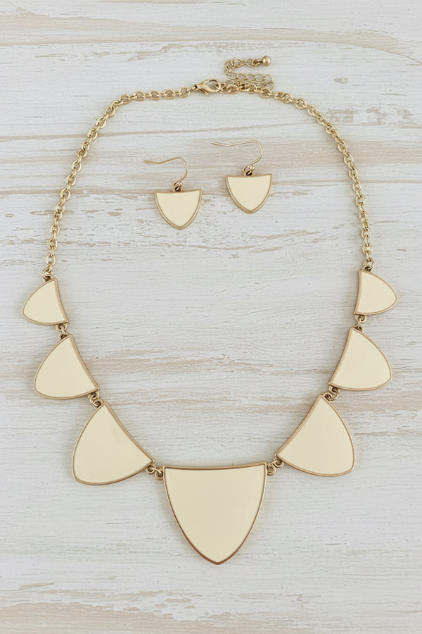 A gold-tone statement necklace and matching drop earrings with shield-shaped cream enamel pendants, displayed on a dark gray stone surface.