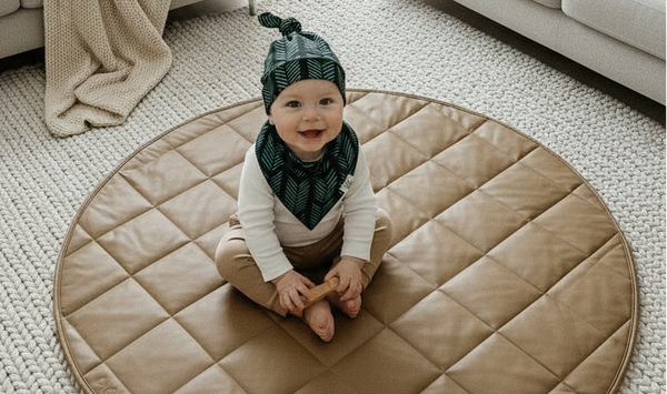 Baby sitting on a round beige mat in a living room with light gray sofas and a lamp.