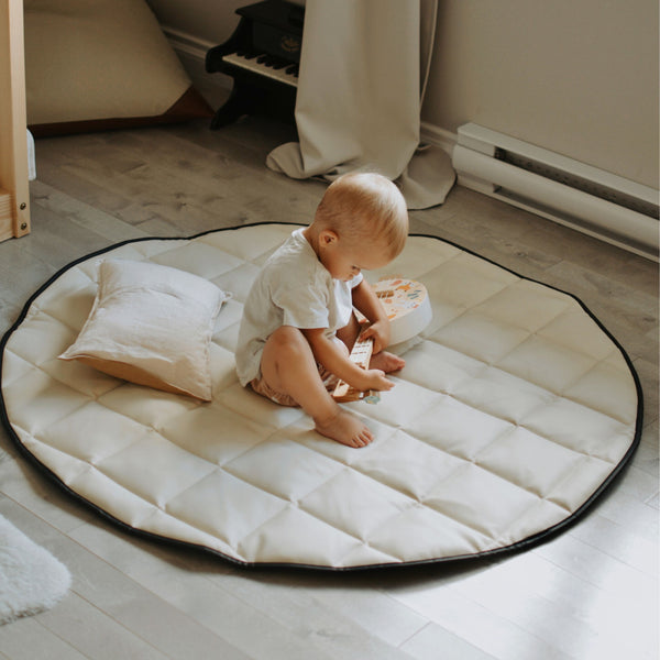 A baby sitting on a round, quilted, black and cream colored padded floor mat with a pillow.