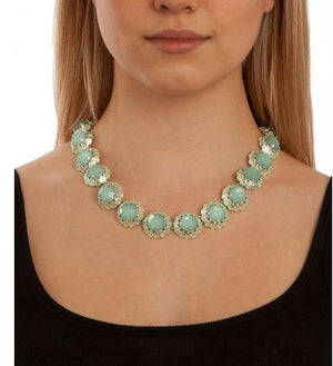 A close-up of a model wearing a chunky statement necklace with light green oval stones in textured gold settings, paired with a black scoop-neck top against a white background.