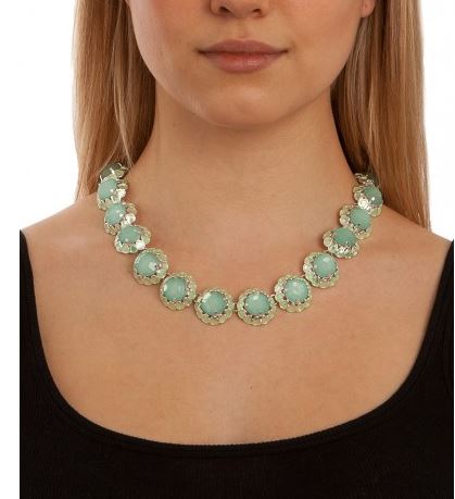 A close-up of a model wearing a chunky statement necklace with light green oval stones in textured gold settings, paired with a black scoop-neck top against a white background.