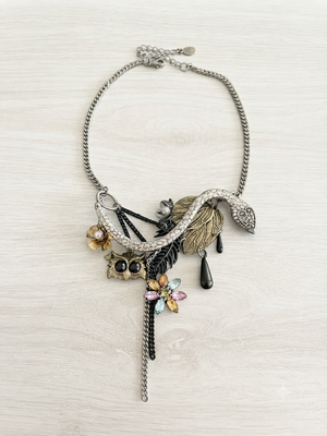 A silver-toned statement necklace featuring a snake, leaf, owl, dragonfly, and teardrop charms, displayed on a light wood background.