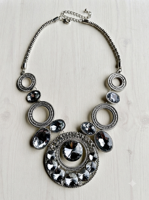 A statement bib necklace featuring a silver-tone chain and multiple circular and oval links adorned with faceted clear and dark gray rhinestones, displayed on a light wood background.