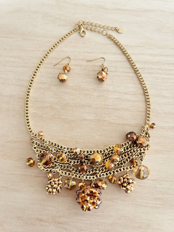 A gold-tone statement necklace with matching drop earrings, featuring multiple layered chains, faceted beads, and dangling crystal-like charms in amber, brown, and gold tones, displayed on a light wood surface.