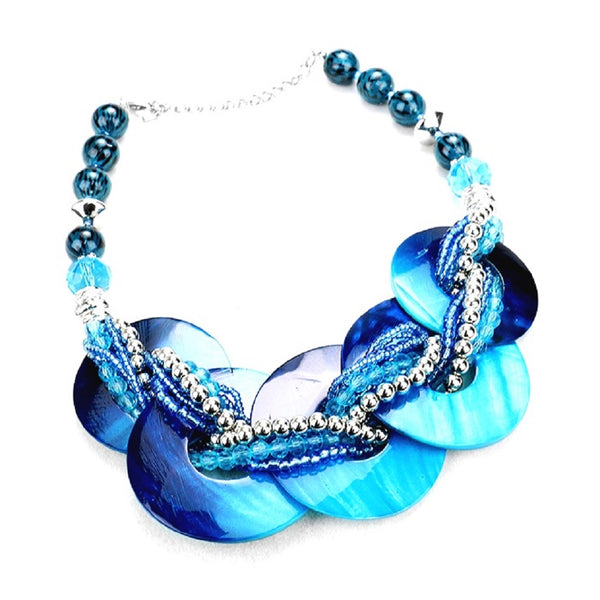 A blue and turquoise statement necklace featuring large iridescent shell-like discs, braided beads, and a silver-tone chain extender on a white background.