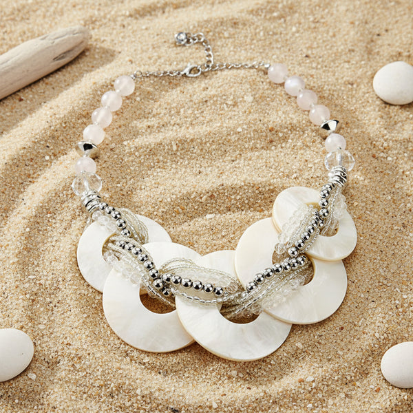 A statement necklace featuring large white mother-of-pearl disc pendants, layered with silver beaded chains, clear crystal beads, and a strand of pale pink pearls on a silver link chain, set against a light gray background.
