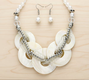 A statement necklace and matching drop earrings set. The necklace features large, overlapping round shell discs in an off-white/cream tone, woven with clear textured cords and accented by silver-tone beaded chains and beads.