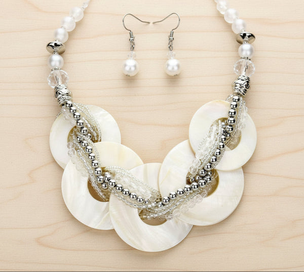 A statement necklace and matching drop earrings set. The necklace features large, overlapping round shell discs in an off-white/cream tone, woven with clear textured cords and accented by silver-tone beaded chains and beads.