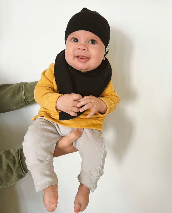 A baby wearing a black bandana bib and a yellow top, sitting and smiling.