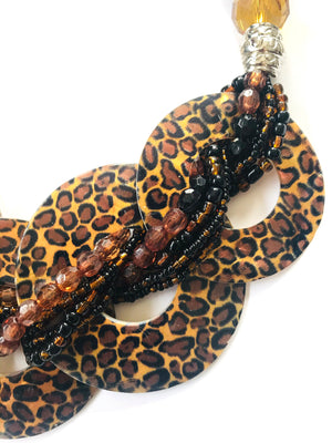 A close-up of a beaded necklace featuring large interlocking circular disc charms in a brown and black leopard print, layered with multi-strand beads in black, brown, and amber.