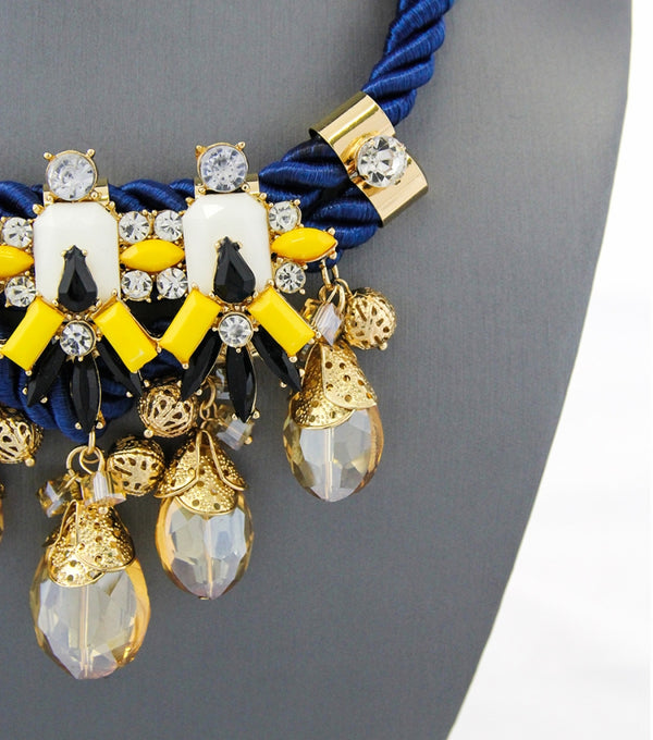 A close-up of a statement bib necklace displayed on a dark gray mannequin bust. The necklace features a royal blue braided cord base with gold-tone metal accents, white rectangular inlays, black teardrop crystals, and dangling faceted clear glass beads with gold caps.