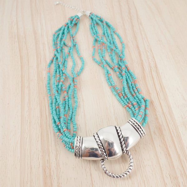 A multi-strand beaded necklace featuring turquoise blue beads with small coral accents, accented by a polished silver-toned cylindrical clasp and a hanging ring, shown on a plain white background.