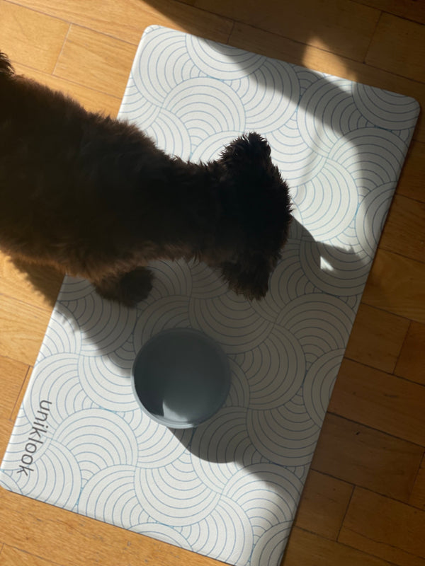 Cat interacting with a gray bowl on a white mat with black patterns on a wooden floor.