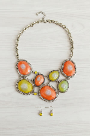 A statement necklace with large orange and yellow agate-like stones in silver-toned settings, shown with matching small yellow dangle earrings on a dark textured surface.