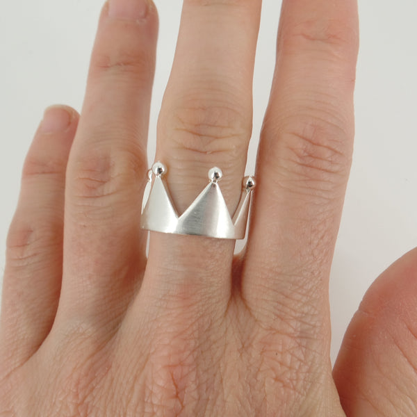 Silver crown-shaped ring on a finger against a plain background
