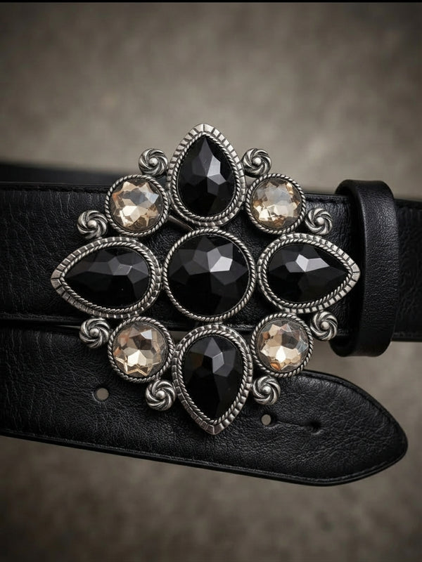 A close-up of a black leather belt featuring an ornate silver-tone buckle adorned with faceted black and smoky-tinted crystals on a textured gray background.
