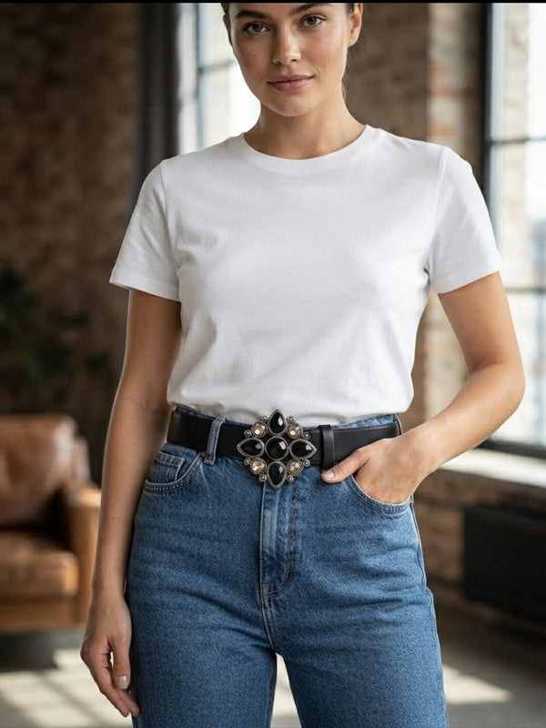 A woman wearing a plain white short-sleeve crewneck t-shirt tucked into blue jeans with a black belt featuring a large decorative silver-toned buckle, standing in a loft-style room with brick walls and windows.