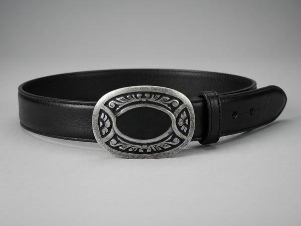 A black leather belt with a large oval silver-tone buckle featuring an engraved floral pattern and a black center, displayed on a neutral gray background.