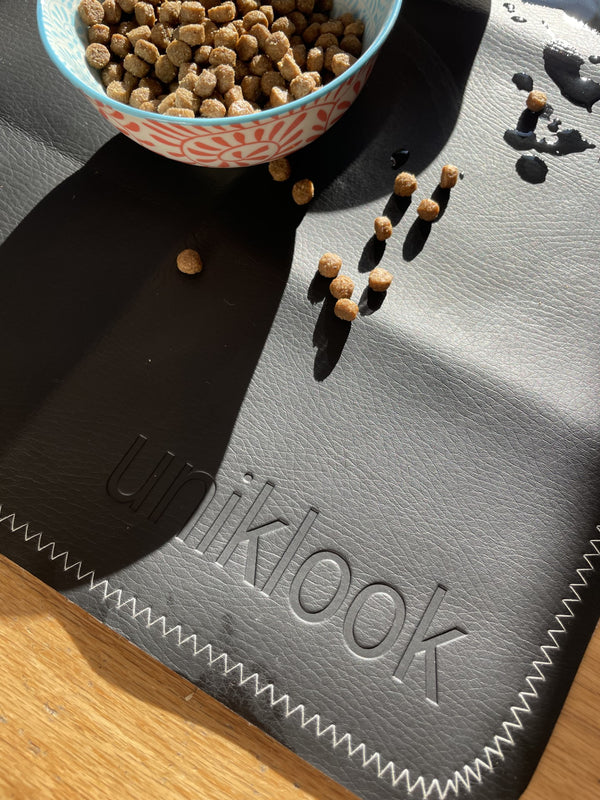 A black and cream vegan leather pet food mat with the brand name 'uniklook' embossed on it, featuring a bowl of cat food on top and a few scattered kibbles around.