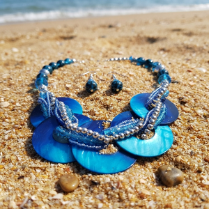 Blue beaded necklace and matching drop earrings displayed on a sandy beach with the ocean in the background.