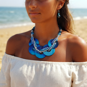 A woman on a beach wearing a chunky blue disc necklace with silver beaded accents and matching blue drop earrings, paired with a white off-the-shoulder top.