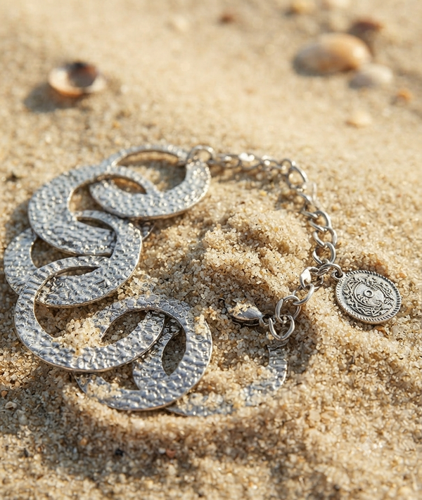 A silver-toned textured bracelet with interlocking circular links and a coin charm, displayed on sandy beach ground.