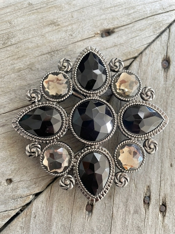 A vintage-style brooch featuring a silver-toned filigree frame with faceted dark smoky gray teardrop and oval stones, accented by smaller champagne-colored round stones, displayed on a rustic weathered wood background.