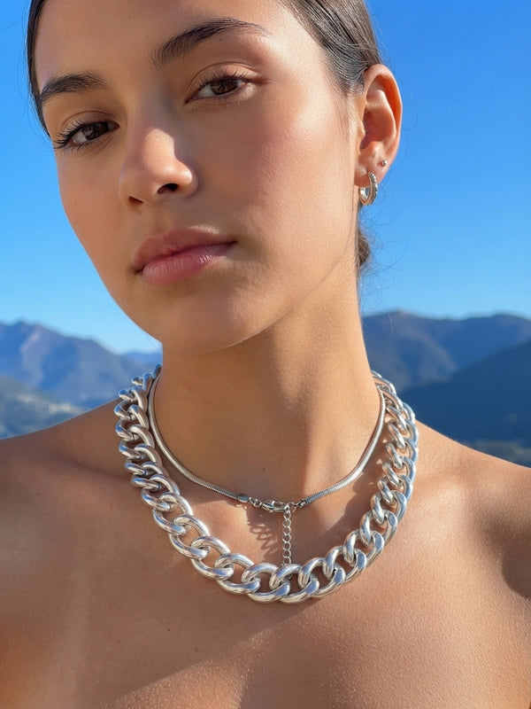 A close-up of a woman wearing a chunky silver chain necklace with a delicate silver pendant, set against a background of blue sky and distant mountains.