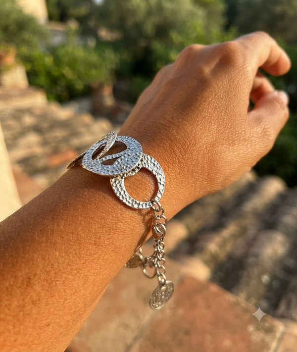 A silver-toned textured chain bracelet with circular links and dangling charms, shown worn on a person's wrist against a blurred outdoor background.