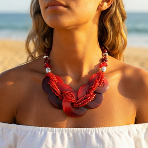 A close-up lifestyle shot of a woman on a beach wearing a chunky statement necklace. The necklace features large, layered translucent red disc elements woven with red beads and accented with small silver beads.
