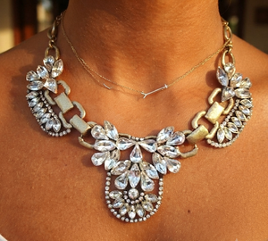 A close-up of a model wearing a statement bib necklace with large clear rhinestones and gold-tone chain links, layered over a delicate gold chain.