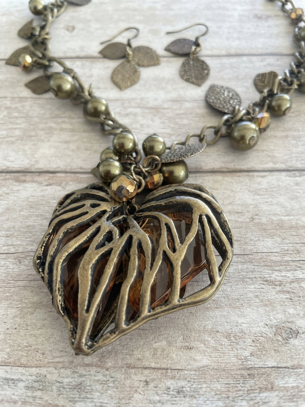 An antique bronze and gold-toned necklace with a large, organic leaf-shaped pendant featuring cutout branches, accompanied by matching dangling leaf earrings, displayed on a light wood background.