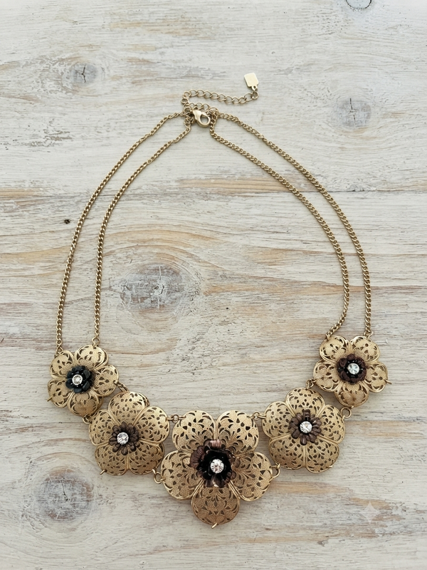 A gold-toned statement necklace featuring five ornate floral elements with dark centers and small clear rhinestones, displayed on a light wood background.