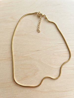 A gold snake chain necklace with an adjustable lobster clasp and extender chain, displayed on a light wood grain surface.