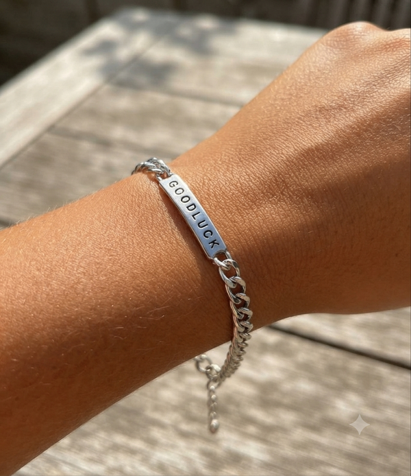 A silver chain bracelet with a polished bar engraved with the word 'GOODLUCK' in black capital letters, shown worn on a person's wrist against a blurred outdoor wooden background.