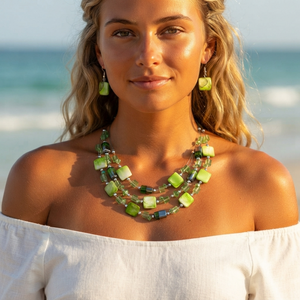 A woman on a beach wearing a green beaded multi-strand necklace and matching drop earrings, with ocean waves blurred in the background.
