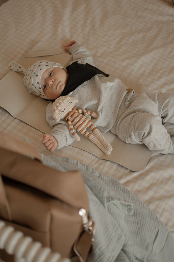 Baby lying on a bed with a striped toy, wearing a hat and holding a brown bag.