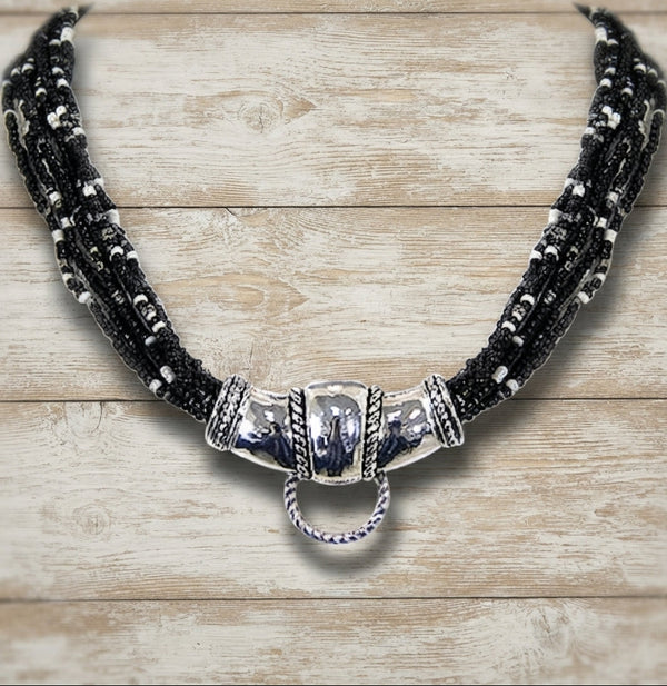 A multi-strand black beaded necklace with a polished silver-toned central clasp and hanging ring, displayed on a light wood plank background.